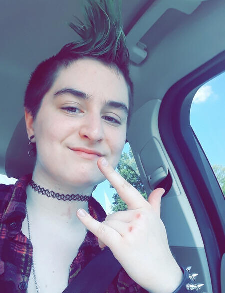 thin white person with a teal mohawk, smiling, giving a devil horns sign with their hand.